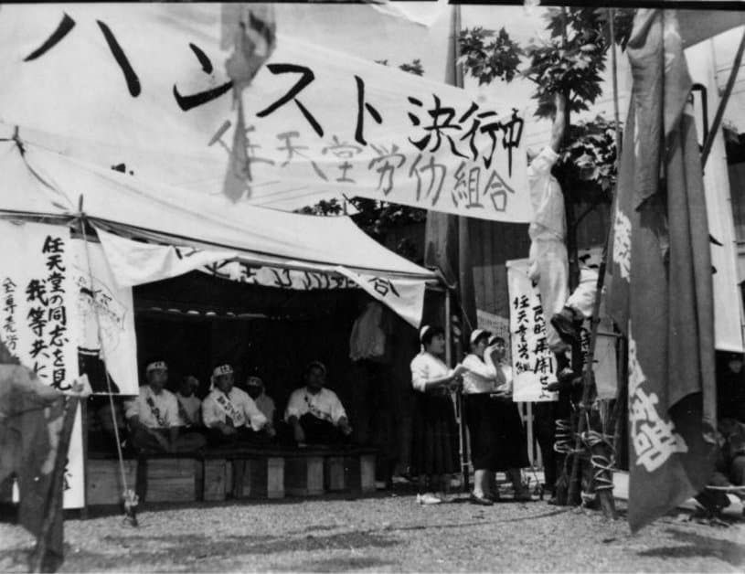 A photo from May 25, 1955. Workers carry a banner which reads, in Japanese, “On A Hunger Strike.” Below that, it reads, “Nintendo Labor Union.”